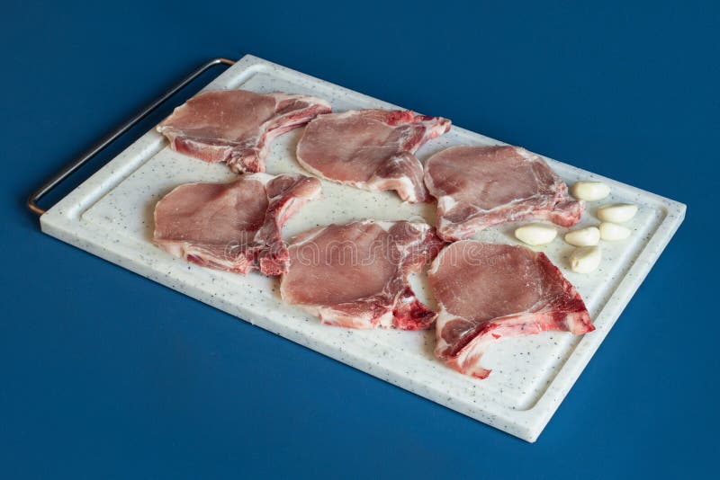 Schweineleitchops mit einer Handvoll Knoblauchzehen auf einem Schnittbrett über einem farbenfrohen, blauen Studiohintergrund zum K stockfotografie
