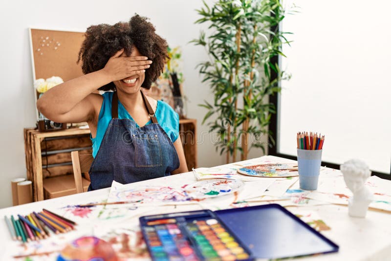 Beautiful african american woman with afro hair painting at art studio smiling and laughing with hand on face covering eyes for surprise. blind concept. Beautiful african american woman with afro hair painting at art studio smiling and laughing with hand on face covering eyes for surprise. blind concept