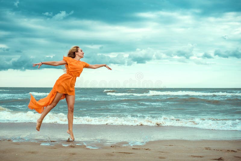 Schöne blonde Frau im orange Minikleid mit dem Fliegenzug, der barfuß auf den nassen Sand in dem stürmenden Meer tanzt lizenzfreies stockfoto Schöne blonde Frau im orange Minikleid mit dem Fliegenzug, der barfuß auf den nassen Sand in dem stürmenden Meer tanzt lizenzfreies stockfoto