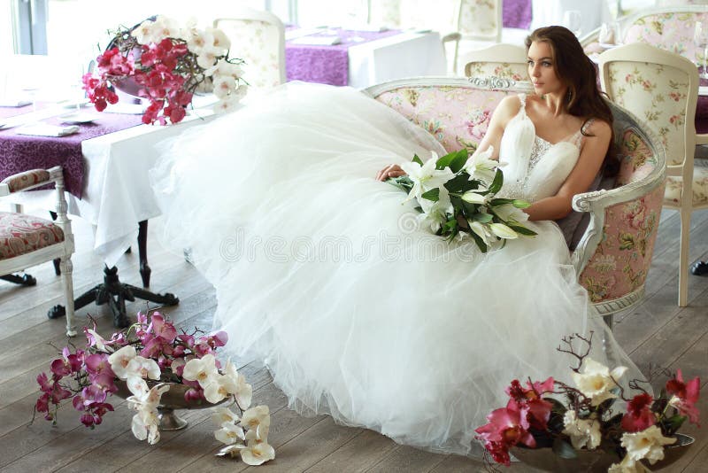 Schöne Braut in einem ausgezeichneten weißen Hochzeitskleid von Tulle mit einem Korsett, das auf dem Sofa mit Blumenstraußlilie u stockfotografie