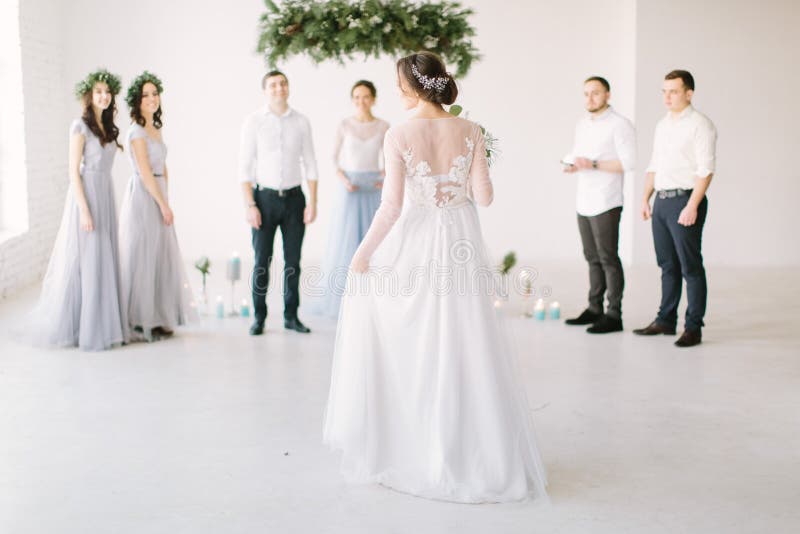 Schöne Braut in einem weißen Kleid hält ein Hochzeitsbrot und geht zu ihrem Bräutigam mit Brautmädchen und Grommern für stockfotografie