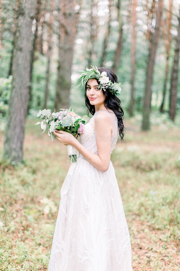 Schöne Braut mit Blumenstrauß in weißem Kleid steht auf Waldboden Rustikaler Stil lizenzfreie stockfotos
