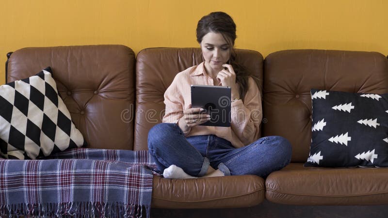 Schöne Brunette-Frau, die Tablette benutzt, während sie auf der Couch zu Hause sitzt Bestandsaufnahmen Websurfen von Frauen, sozi