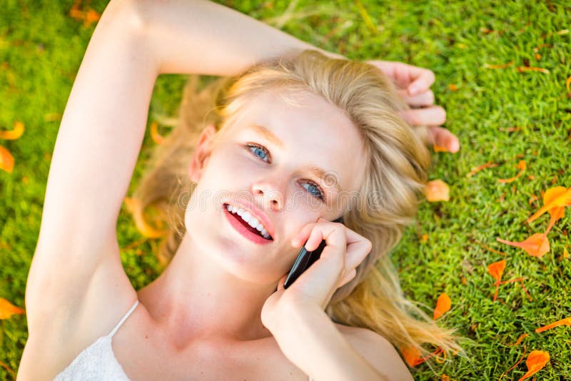 Schöne glückliche Frau, die um ein Mobiltelefon beim Lügen auf dem grünen Gras während der Herbstzeit ersucht lizenzfreie stockfotos Schöne glückliche Frau, die um ein Mobiltelefon beim Lügen auf dem grünen Gras während der Herbstzeit ersucht lizenzfreie stockfotos