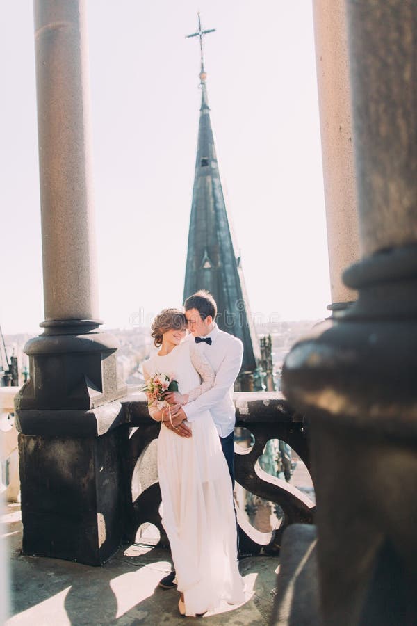 Schöne Hochzeitspaarjungvermählten, die auf Balkon mit Ansicht der alten Stadt umarmen lizenzfreie stockfotografie Schöne Hochzeitspaarjungvermählten, die auf Balkon mit Ansicht der alten Stadt umarmen lizenzfreie stockfotografie