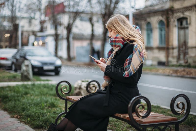 Schöne junge Frau des Porträts auf Bank im städtischen Hintergrund, der Telefon schaut lizenzfreie stockbilder Schöne junge Frau des Porträts auf Bank im städtischen Hintergrund, der Telefon schaut lizenzfreie stockbilder