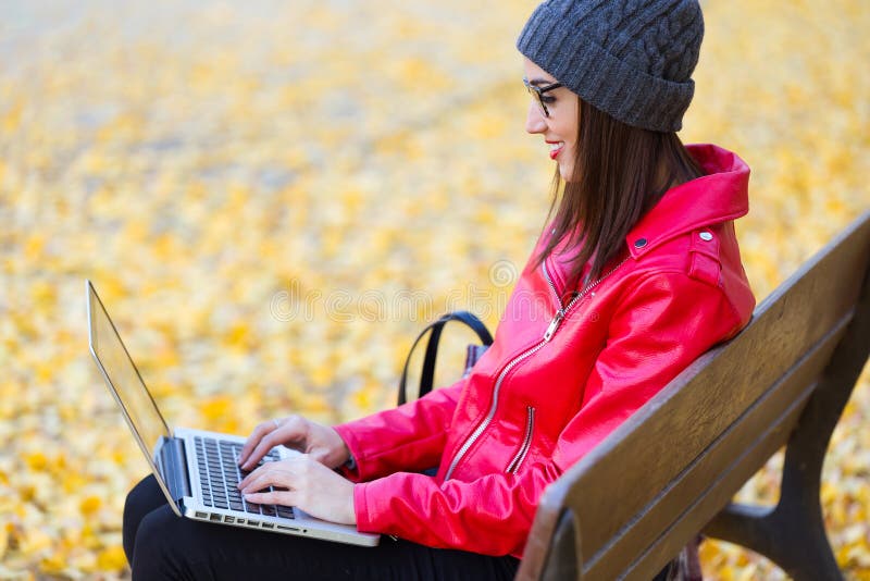 Schöne junge Frau, die in einer Bank sitzt und ihren Laptop im Herbst verwendet stockfotografie Schöne junge Frau, die in einer Bank sitzt und ihren Laptop im Herbst verwendet stockfotografie