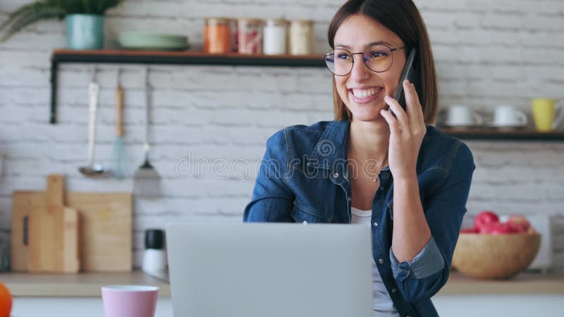 Schöne junge Geschäftsfrau, die mit ihrem Handy und mit Computer in der Küche zu Hause arbeiten wird.