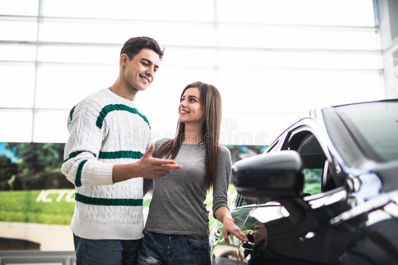 Schöne junge Paare, die an der Verkaufsstelle beschließt das Auto, um zu kaufen stehen Mann gezeigt auf Auto lizenzfreies stockbild Schöne junge Paare, die an der Verkaufsstelle beschließt das Auto, um zu kaufen stehen Mann gezeigt auf Auto lizenzfreies stockbild