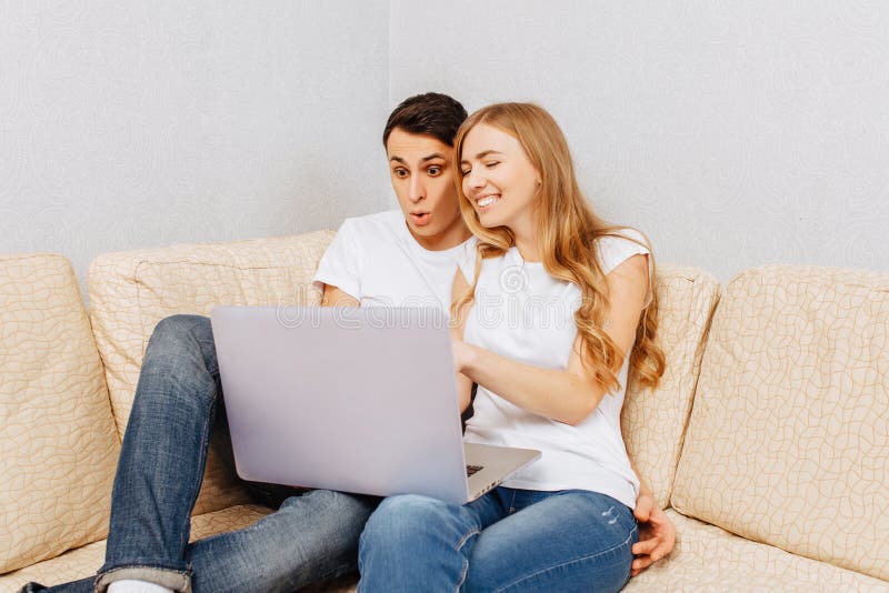 Schöne junge Paare, Mann und Frau, die einen Laptop, das Umarmen und das Lächeln, zu Hause sitzend auf der Couch verwendet stockbild Schöne junge Paare, Mann und Frau, die einen Laptop, das Umarmen und das Lächeln, zu Hause sitzend auf der Couch verwendet stockbild