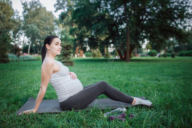 Schöne junge schwangere Frau auf Yogakameraden im Park draußen sitzen Sie hält eine Hand auf Bauch und Mageres auf anderen Frau stockfotos Schöne junge schwangere Frau auf Yogakameraden im Park draußen sitzen Sie hält eine Hand auf Bauch und Mageres auf anderen Frau stockfotos