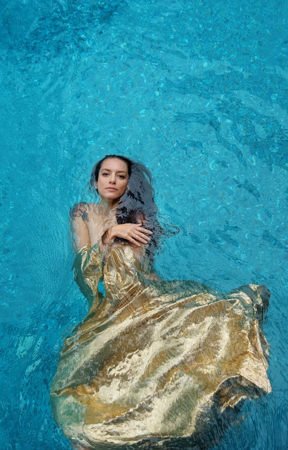 Schöne junge stolze Frau im goldenen Kleid, Abendkleid, welches das leicht elegante Schwimmen in das Wasser im Pool dunkel schwim lizenzfreie stockbilder
