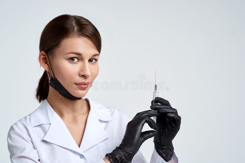 Schöne junge Ärztin in der medizinischen Robe, in der Schutzmaske und in den medizinischen schwarzen Handschuhen hält Spritze mit lizenzfreie stockbilder Schöne junge Ärztin in der medizinischen Robe, in der Schutzmaske und in den medizinischen schwarzen Handschuhen hält Spritze mit lizenzfreie stockbilder