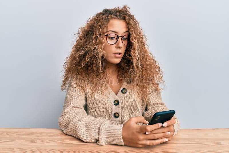 Beautiful caucasian teenager girl using smartphone sitting on the table clueless and confused expression. doubt concept. Beautiful caucasian teenager girl using smartphone sitting on the table clueless and confused expression. doubt concept