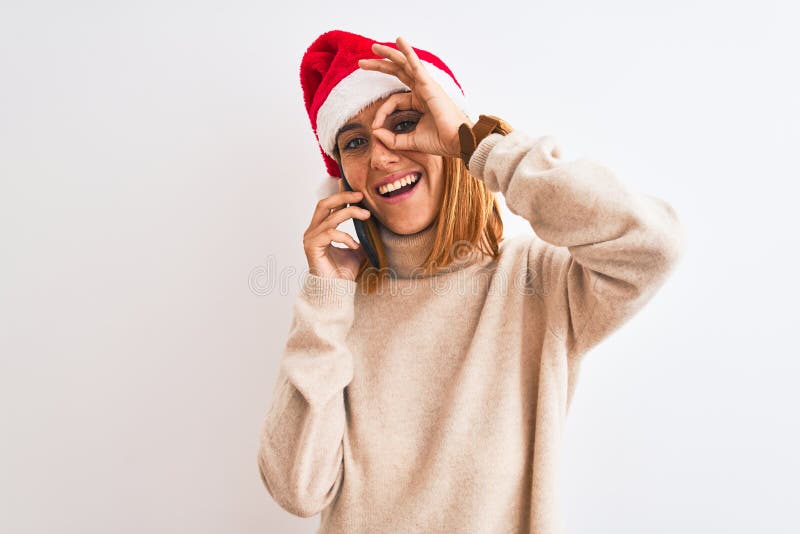 Schöne Rotkopfdame mit Weihnachtsmütze, die auf Smartphone mit fröhlichem Gesicht spricht und mit gutem Auge ein Zeichen mit Hand lizenzfreie stockbilder