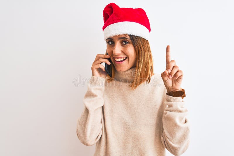 Schöne Rotkopfdame mit Weihnachtsmütze, die auf Smartphone spricht, überrascht mit einer Idee oder einem Fragefinger mit fröhliche lizenzfreie stockbilder