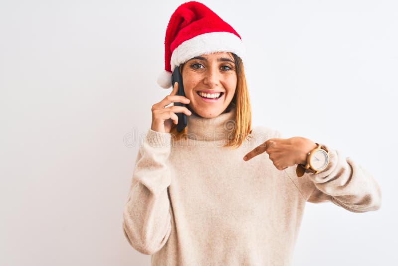 Schöne Rotkopffrau mit Weihnachtsmütze, die auf dem Smartphone mit Überraschungsgesicht auf sich selbst zeigt stockbilder