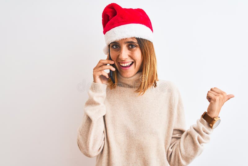 Schöne Rotkopffrau mit Weihnachtsmütze, die auf Smartphone zeigt und mit Daumen zur Seite zeigt, mit glücklichem Gesicht stockbilder