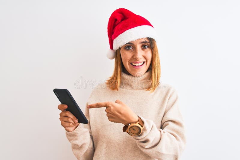 Schöne Rotkopffrau mit Weihnachtsmütze und Smartphone sehr glücklich mit Hand und Finger zeigen stockfotografie