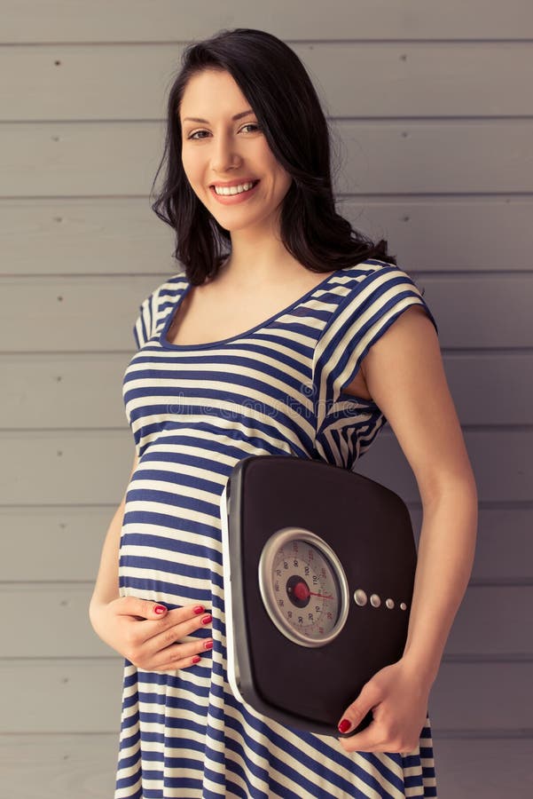 Beautiful pregnant woman is holding weigh scales, keeping one hand on a belly, looking at camera and smiling. Beautiful pregnant woman is holding weigh scales, keeping one hand on a belly, looking at camera and smiling