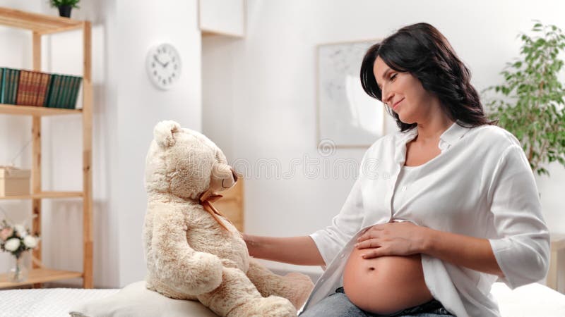 Schöne schwangere Frau, die Bärenspielzeug berührt, wartet Baby mit Liebe Weiße Zimmer in der Nähe