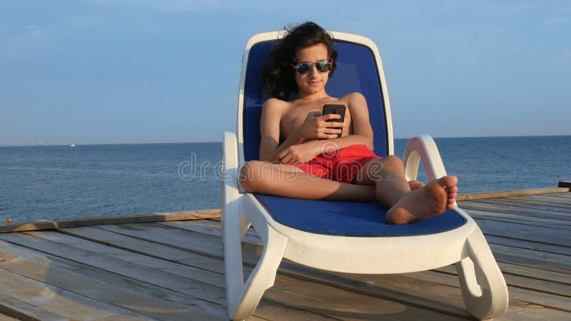 Schöner Jugendlichjunge mit gelockten Lügen des schwarzen Haares auf einem Klappstuhl auf einer hölzernen Terrasse über dem Meer