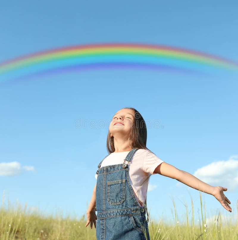 Schöner Regenbogen in blauem Himmel über süßes kleines Mädchen auf dem Feld an sonnigen Tag stockfoto