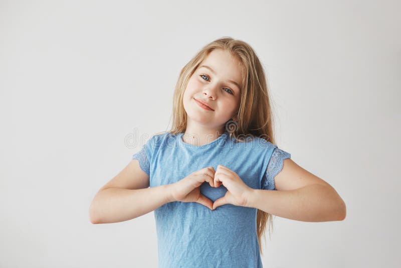 Schönes blondes Mädchen mit dem hellen Haar im blauen T-Shirt, das in camera mit dem leichten Lächeln, Herzgeste mit machend scha