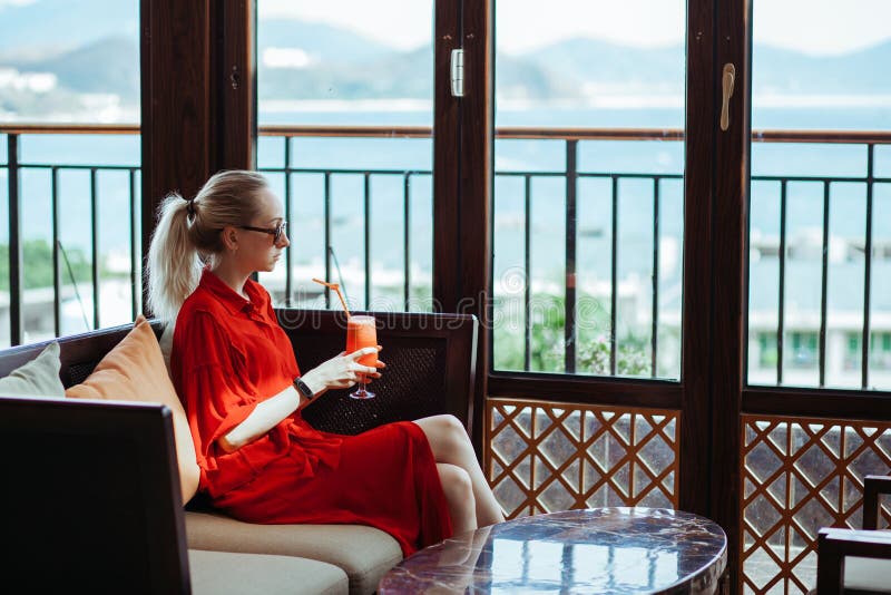 Schönes junges blondes Mädchen im roten Kleid und in der Sonnenbrille rotes Cocktail von einem Glas auf einer Freiterrasse von tr lizenzfreies stockbild Schönes junges blondes Mädchen im roten Kleid und in der Sonnenbrille rotes Cocktail von einem Glas auf einer Freiterrasse von tr lizenzfreies stockbild