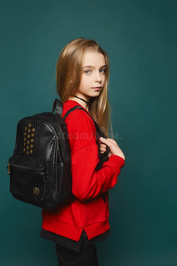 Beautiful young model teen girl with long blonde hair in red sweatshirt and with backpack posing at the dark-green background. Beautiful young model teen girl with long blonde hair in red sweatshirt and with backpack posing at the dark-green background.