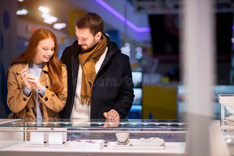 Newly married couple, caucasian men and women during shopping in mall, choose expensive jewelry for female, wearing coat and hugging man. Newly married couple, caucasian men and women during shopping in mall, choose expensive jewelry for female, wearing coat and hugging man