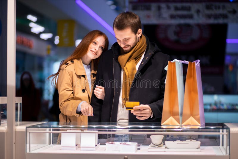 Newly married couple, caucasian men and women during shopping in mall, choose expensive jewelry for female, wearing coat and hugging man. Newly married couple, caucasian men and women during shopping in mall, choose expensive jewelry for female, wearing coat and hugging man