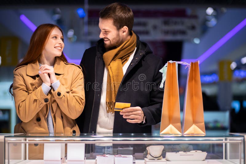 Newly married couple, caucasian men and women during shopping in mall, choose expensive jewelry for female, wearing coat and hugging man. Newly married couple, caucasian men and women during shopping in mall, choose expensive jewelry for female, wearing coat and hugging man