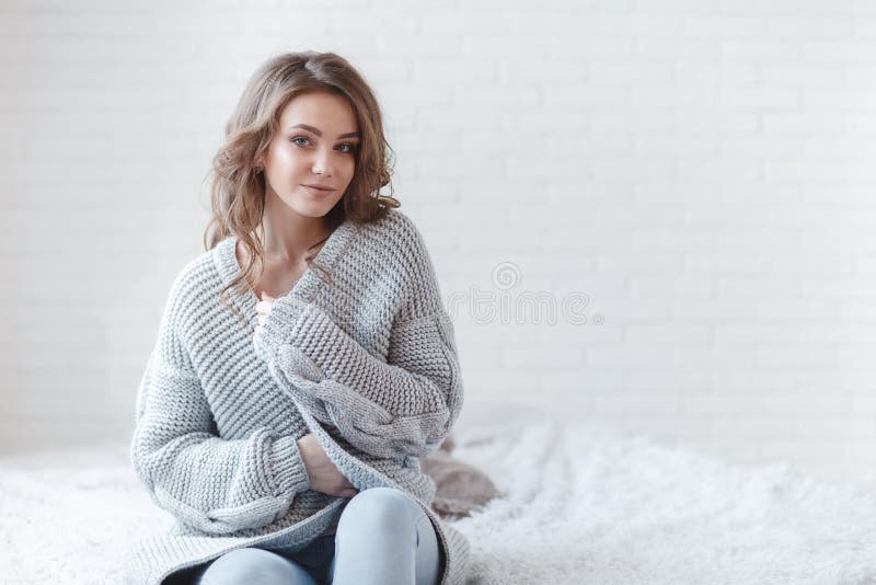 Winter Concept. Cute young girl in a gray knitted sweater. Beautiful woman is relaxing in a white bedroom. Beautiful woman in winter clothes are waking up in the morning. Woman wearing a sweater in a white bedroom. Winter Concept. Cute young girl in a gray knitted sweater. Beautiful woman is relaxing in a white bedroom. Beautiful woman in winter clothes are waking up in the morning. Woman wearing a sweater in a white bedroom.