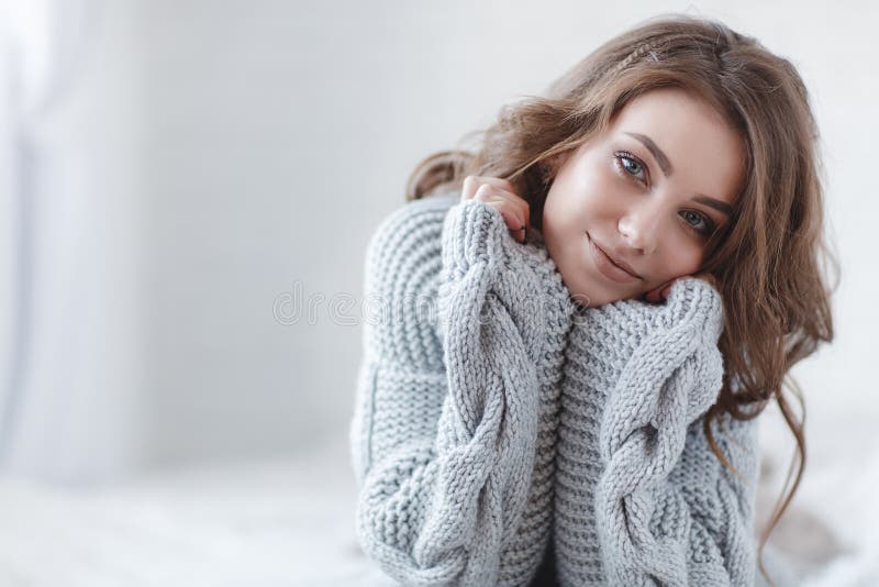 Winter Concept. Cute young girl in a gray knitted sweater. Beautiful woman is relaxing in a white bedroom. Beautiful woman in winter clothes are waking up in the morning. Woman wearing a sweater in a white bedroom. Winter Concept. Cute young girl in a gray knitted sweater. Beautiful woman is relaxing in a white bedroom. Beautiful woman in winter clothes are waking up in the morning. Woman wearing a sweater in a white bedroom.