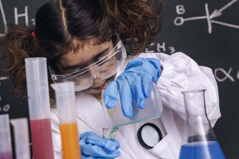 Little scientist girl with goggles and gloves in lab coat mixing chemical liquids in flasks, blackboard background with science formulas, explosion in the laboratory, back to school concept. Little scientist girl with goggles and gloves in lab coat mixing chemical liquids in flasks, blackboard background with science formulas, explosion in the laboratory, back to school concept