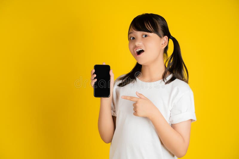 Scioccata ragazzina con una maglietta bianca che mostra uno smartphone con uno schermo bianco su uno sfondo giallo. tecnologia del immagine stock libera da diritti