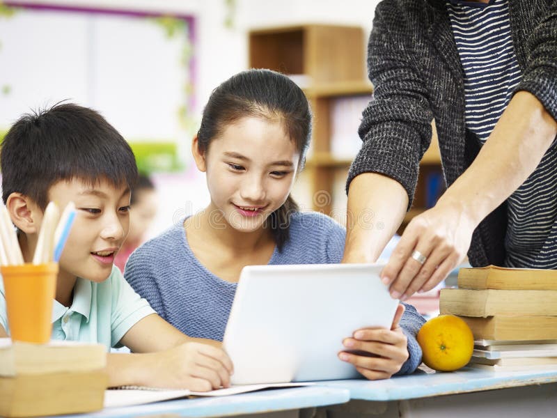 Two asian elementary school children using digital tablet with help from teacher tutor in classroom. Two asian elementary school children using digital tablet with help from teacher tutor in classroom.