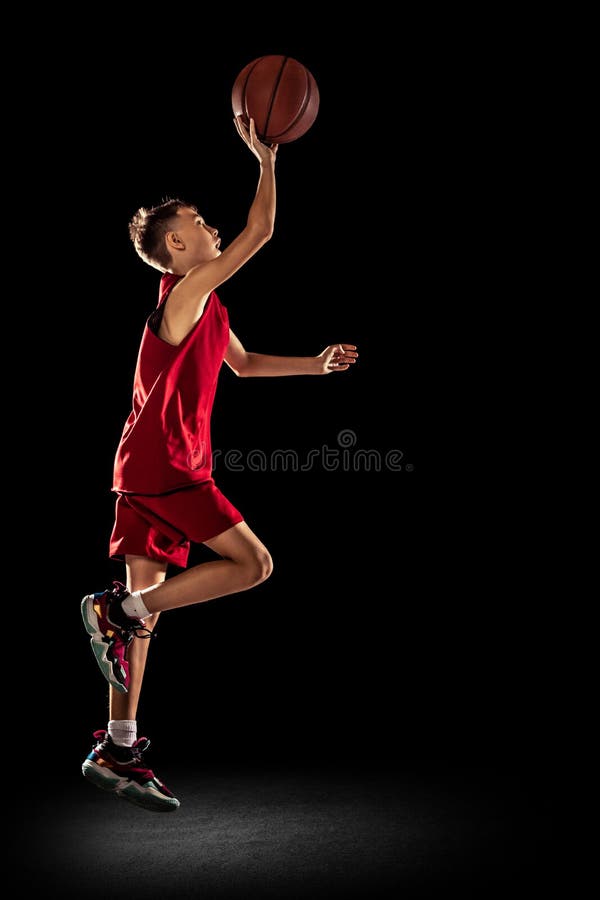 Scoring goal into jump. Sportive teen boy, basketball player throwing ball into the basket isolated over black