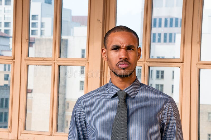 A well dressed, young African American male standing in front of a window with office buildings in the background scowls in anger. A well dressed, young African American male standing in front of a window with office buildings in the background scowls in anger.