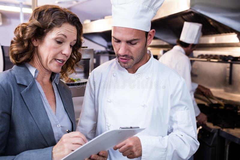 Scrittura femminile del responsabile del ristorante sulla lavagna per appunti mentre interagendo al cuoco unico capo immagine stock Scrittura femminile del responsabile del ristorante sulla lavagna per appunti mentre interagendo al cuoco unico capo immagine stock