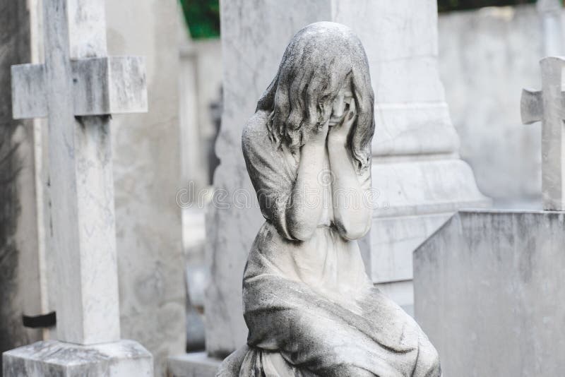 Sculpture of a angel girl  in an old cemetery. Closeup of stoned angel with closed eyes and cross monument at cemetery. Graveyard