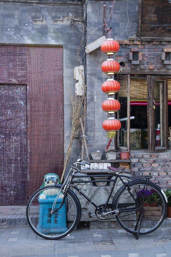 A homemade broom, an overflowing blue garbage bin with Chinese lettering, some flowerpots, a bicycle,and a red Chinese lantern on gray pavement in front of a rust colored closed-up doorway next to an open window make up this local Chinese street scene, or Hutong, in old Beijing. A homemade broom, an overflowing blue garbage bin with Chinese lettering, some flowerpots, a bicycle,and a red Chinese lantern on gray pavement in front of a rust colored closed-up doorway next to an open window make up this local Chinese street scene, or Hutong, in old Beijing.