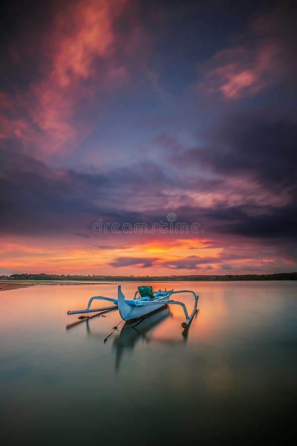 Seascape. Fisherman boat. Traditional Balinese boat jukung. Fishing boat at the beach during sunrise. Water reflection. Cloudy sky. Sunlight on horizon. Slow