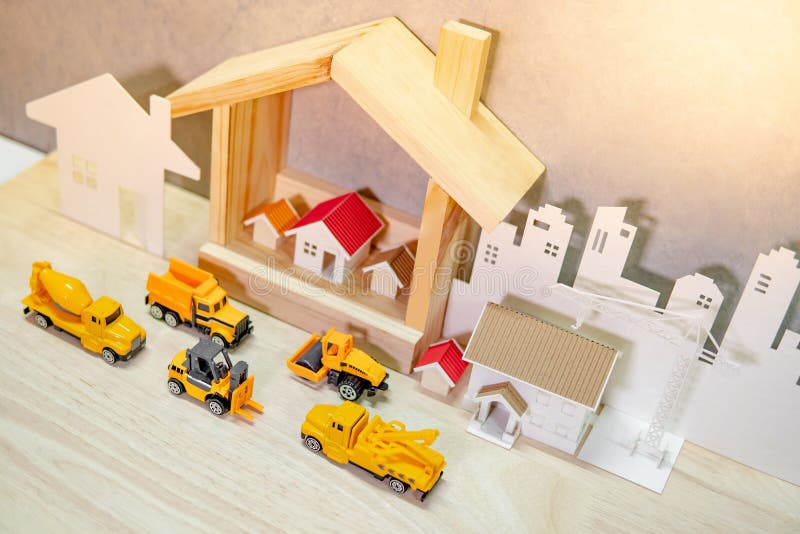 House models, paper crane and city background with miniature yellow trucks on wooden table. Architecture and construction industry for housing development business. Property or real estate concept. House models, paper crane and city background with miniature yellow trucks on wooden table. Architecture and construction industry for housing development business. Property or real estate concept
