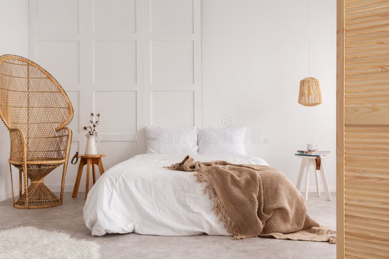 Sedia del rattan e tavola di legno accanto al letto con la coperta marrone nell'interno bianco della camera da letto Foto reale fotografie stock libere da diritti