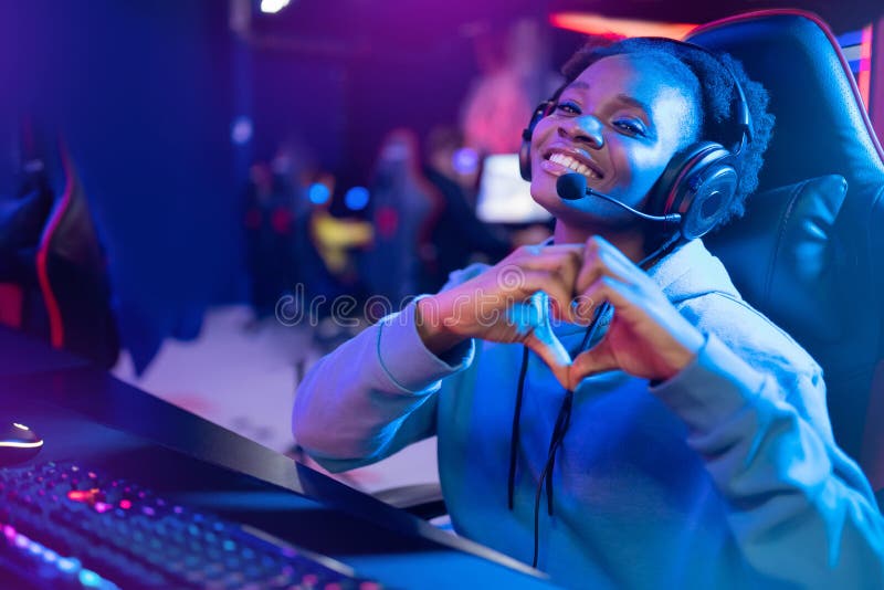 Seducente ragazza africana mostra l'insegna del cuore con un giocatore professionista che gioca online videogame a colori fotografia stock