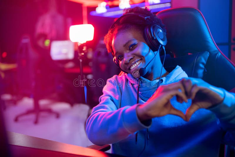 Seducente ragazza afro-americana bella mostra il segno del cuore con un giocatore professionista che gioca con un computer per gio immagini stock