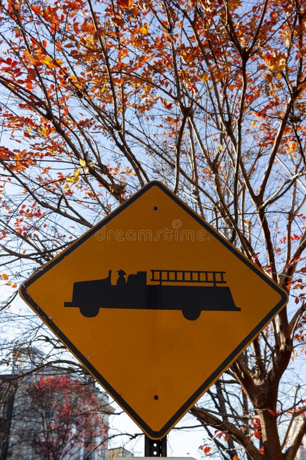 Emergency vehicle traffic sign on side of a road with a vintage fire engine symbol on reflective yellow quadrangular metal post. Background has trees with vibrant autumn leaves and blue sky. Emergency vehicle traffic sign on side of a road with a vintage fire engine symbol on reflective yellow quadrangular metal post. Background has trees with vibrant autumn leaves and blue sky