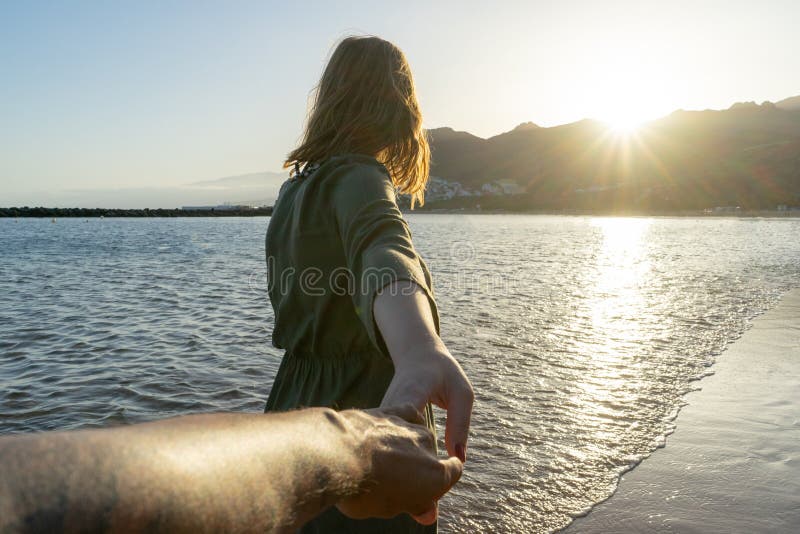 Seguito della fidanzata con un vestito estivo che corre e tiene la mano del fidanzato sulla spiaggia al tramonto Felice coppia in fotografia stock